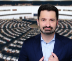 Ein Mann mit kurzem Bart in einem Blauen Anzug vor dem eurpäischen Parlament, das im Hintergrund unscharf dargestellt ist